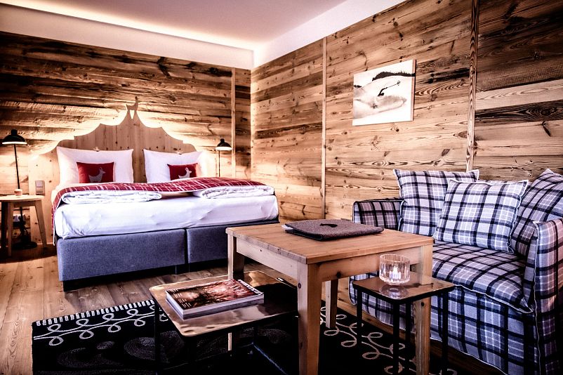 Gemütliches Hotelzimmer mit rustikalen Holzwänden, einem Doppelbett mit roten Kissen, einem karierten Sofa, und einem Holztisch auf einem gemusterten Teppich.