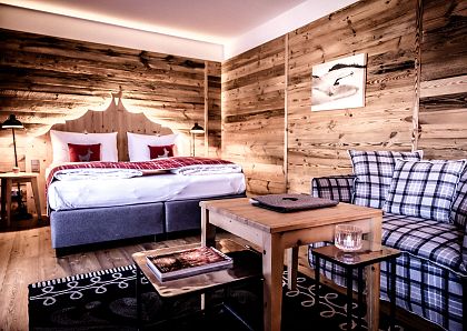Gemütliches Hotelzimmer mit rustikalen Holzwänden, einem Doppelbett mit roten Kissen, einem karierten Sofa, und einem Holztisch auf einem gemusterten Teppich.