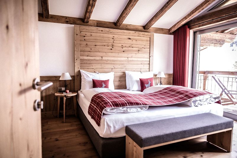 Gemütliches Hotelzimmer mit Holzwänden und freiliegenden Balken, einem großen Bett mit roter Tagesdecke und Kissen, sowie einem Balkon mit Ausblick.