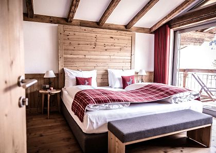 Gemütliches Hotelzimmer mit Holzwänden und freiliegenden Balken, einem großen Bett mit roter Tagesdecke und Kissen, sowie einem Balkon mit Ausblick.