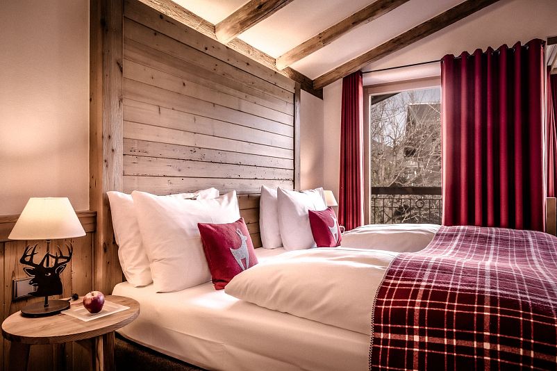 Gemütliches Schlafzimmer im Hüttendesign mit Holzwänden, zwei Einzelbetten, rotkarierten Decken, einem Hirschkissen und warmem, weichem Licht von einer Lampe.
