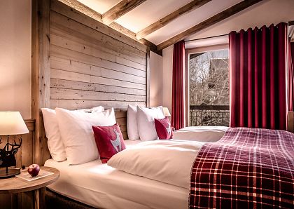 Gemütliches Schlafzimmer im Hüttendesign mit Holzwänden, zwei Einzelbetten, rotkarierten Decken, einem Hirschkissen und warmem, weichem Licht von einer Lampe.