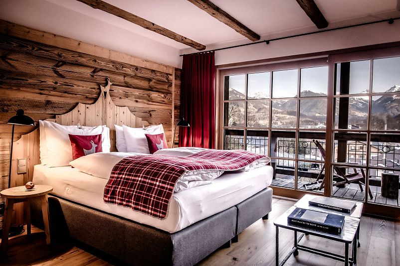Gemütliches Schlafzimmer mit rustikalem Holzdekor, großem Bett und plaidierter Decke. Große Fenster bieten Blick auf die schneebedeckten Berge in der Ferne.