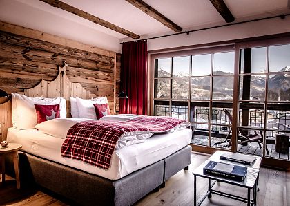 Gemütliches Schlafzimmer mit rustikalem Holzdekor, großem Bett und plaidierter Decke. Große Fenster bieten Blick auf die schneebedeckten Berge in der Ferne.
