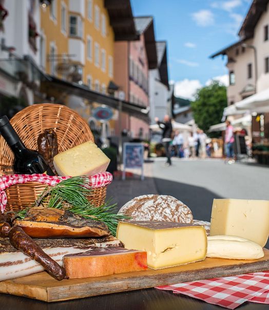 Marktszene auf einer gepflasterten Straße in einem malerischen Dorf. Im Vordergrund ein Tisch mit Käse, Brot und einer Weinflasche. Menschen und Stände im Hintergrund.
