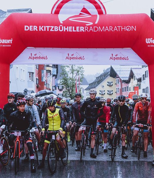 Teilnehmer des Kitzbüheler Radmarathons stehen an der Startlinie unter einem großen roten Torbogen. Viele Radfahrer tragen bunte Sportbekleidung und Helme.