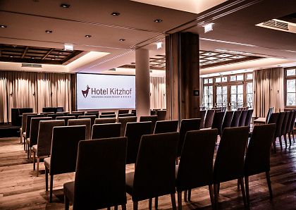 Ein moderner Konferenzraum mit Reihen von gepolsterten Stühlen, einer großen Leinwand vorne, die das Logo des Hotel Kitzhof zeigt, und großen Fenstern rechts.