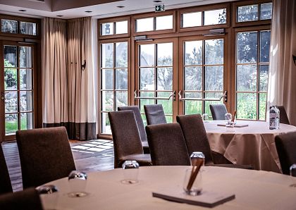 Ein eleganter, heller Konferenzraum mit runden Tischen, gedeckt mit Notizblöcken und Flaschen. Große Fenster lassen viel Tageslicht herein, Blick ins Grüne.