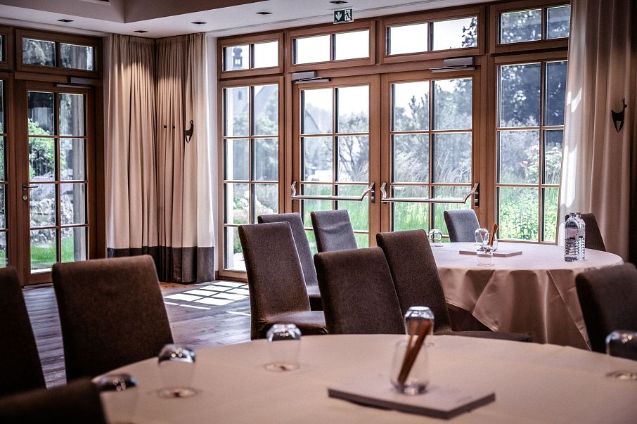 Ein eleganter, heller Konferenzraum mit runden Tischen, gedeckt mit Notizblöcken und Flaschen. Große Fenster lassen viel Tageslicht herein, Blick ins Grüne.