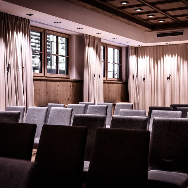Ein leerer Konferenzraum mit grauen Stühlen in Reihen angeordnet. Holzwände umrahmen den Raum, und mehrere Fenster lassen Tageslicht herein.