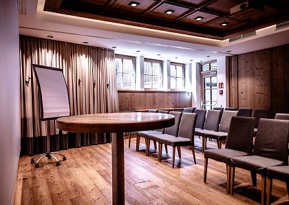 Ein moderner Konferenzraum mit Holzboden und neutralen Farben, ausgestattet mit grauen Stühlen, einem runden Tisch und einem Flipchart vor Vorhängen.