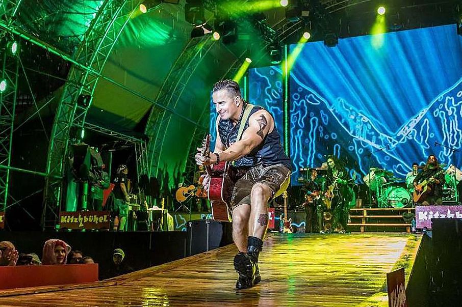 Ein Musiker auf einer Bühne spielt E-Gitarre. Er trägt ein ärmelloses Shirt und Shorts. Hinter ihm leuchtet ein bunter Hintergrund, umgeben von Bandmitgliedern.
