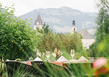 Ein gemütlicher Tisch im Freien mit gedeckten Plätzen und Weingläsern. Im Hintergrund sind grüne Bäume und eine Kirche vor hügeliger Landschaft zu sehen.