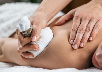 Nahaufnahme einer entspannenden Massage: Eine Person erhält eine Massage mit einem Kräuterstempel. Hände drücken sanft auf den Rücken, um Verspannungen zu lösen.