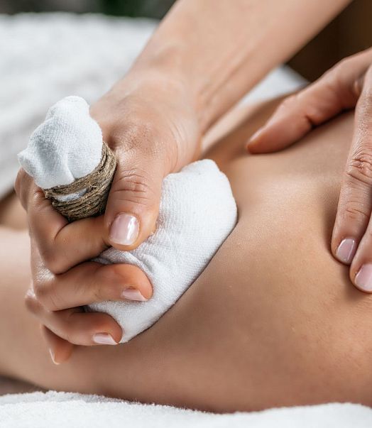 Nahaufnahme einer entspannenden Massage: Eine Person erhält eine Massage mit einem Kräuterstempel. Hände drücken sanft auf den Rücken, um Verspannungen zu lösen.