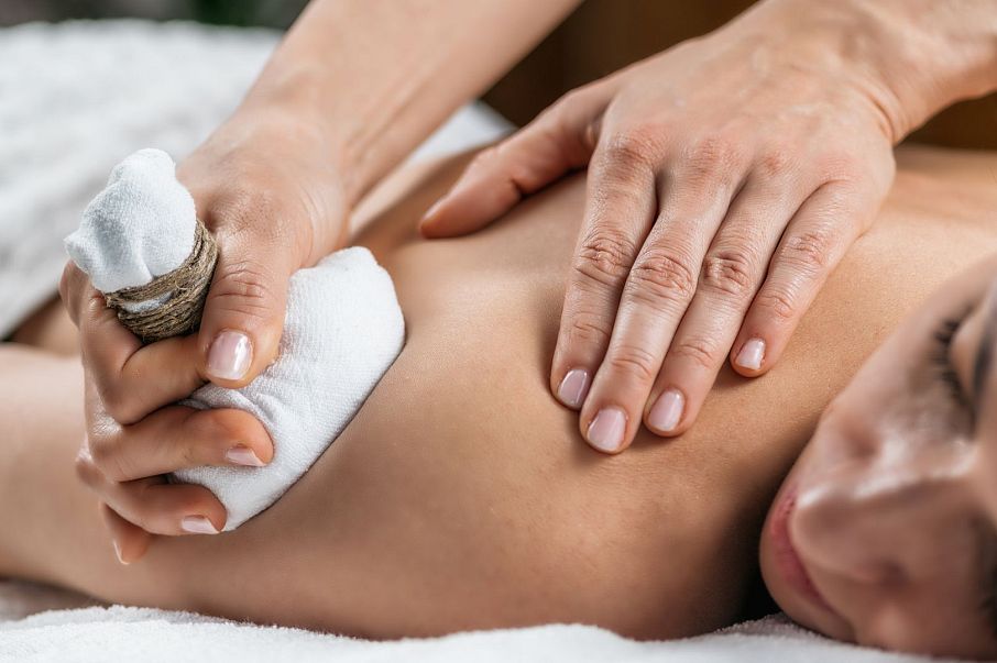 Nahaufnahme einer entspannenden Massage: Eine Person erhält eine Massage mit einem Kräuterstempel. Hände drücken sanft auf den Rücken, um Verspannungen zu lösen.