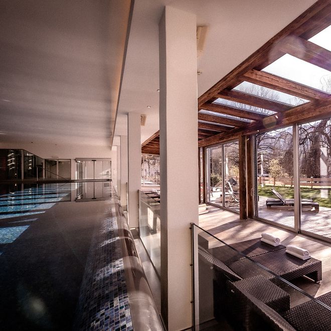 Innenraum eines modernen Spa-Bereichs mit großen Fenstern und Tageslicht. Links ein langer Pool, rechts Liegen mit Handtüchern, dahinter Bäume im Freien.
