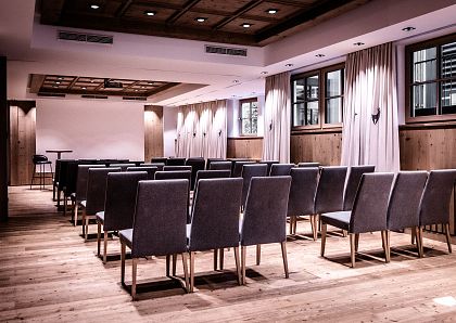 Ein eleganter, moderner Konferenzraum mit Holzboden und -decke. Reihen grauer Stühle sind auf das Präsentationspult und eine leere Leinwand ausgerichtet.
