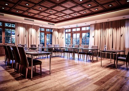 Ein eleganter Konferenzraum mit Holzboden und Holzwandpaneelen. Rechteckige Tische und gepolsterte Stühle sind arrangiert. Große Fenster lassen Tageslicht herein.
