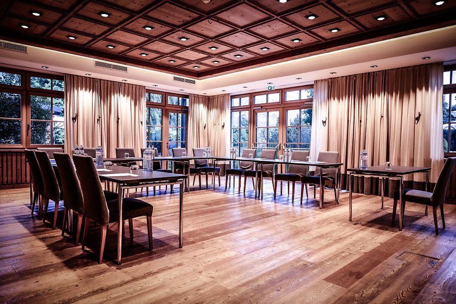 Ein eleganter Konferenzraum mit Holzboden und Holzwandpaneelen. Rechteckige Tische und gepolsterte Stühle sind arrangiert. Große Fenster lassen Tageslicht herein.