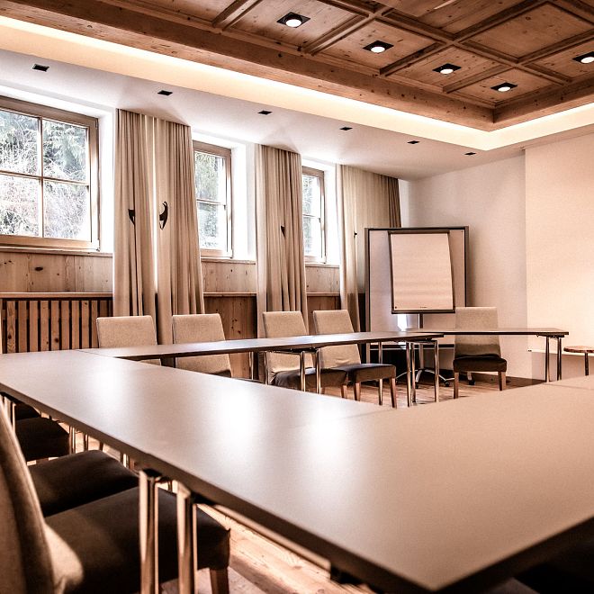 Ein leerer, moderner Konferenzraum mit U-förmiger Anordnung der Tische, umgeben von bequemen Stühlen. An einer Wand steht ein Flipchart, Fenster lassen Licht herein.