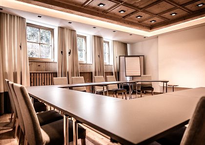 Ein leerer, moderner Konferenzraum mit U-förmiger Anordnung der Tische, umgeben von bequemen Stühlen. An einer Wand steht ein Flipchart, Fenster lassen Licht herein.