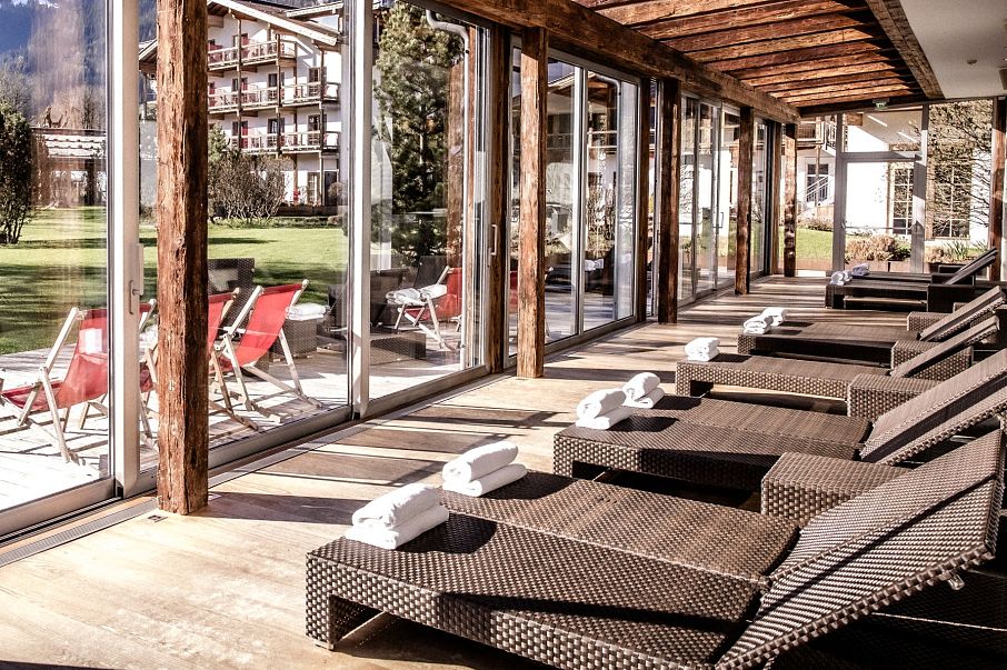 Eine sonnendurchflutete Entspannungslounge mit Liegestühlen aus Rattan und Blick auf einen weitläufigen, gepflegten Garten mit grünen Bäumen und Bergen im Hintergrund.