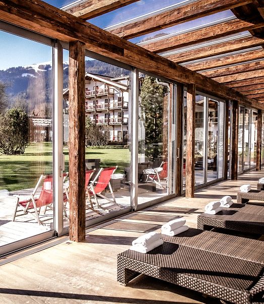 Eine sonnendurchflutete Entspannungslounge mit Liegestühlen aus Rattan und Blick auf einen weitläufigen, gepflegten Garten mit grünen Bäumen und Bergen im Hintergrund.