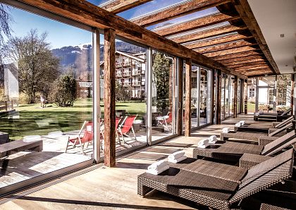 Eine sonnendurchflutete Entspannungslounge mit Liegestühlen aus Rattan und Blick auf einen weitläufigen, gepflegten Garten mit grünen Bäumen und Bergen im Hintergrund.