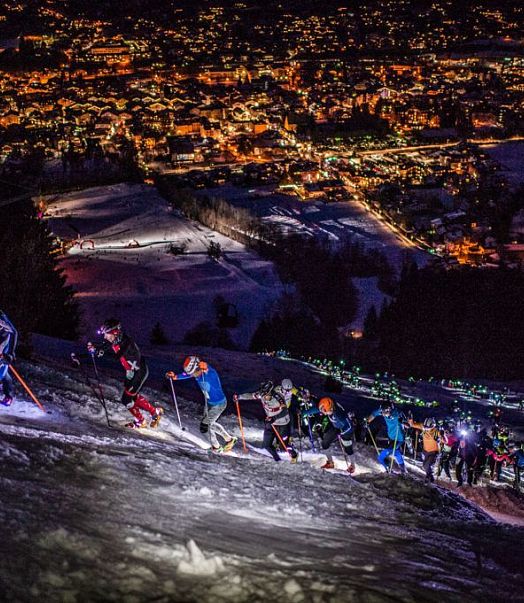 Menschen steigen im Dunkeln mit Stirnlampen auf einem schneebedeckten Bergpfad hinauf, während eine beleuchtete Stadt im Hintergrund zu sehen ist.