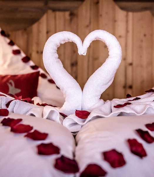 Ein romantisch dekoriertes Bett mit roten Rosenblättern und zwei weißen Handtüchern, die zu einem Herz geformt sind. Der Hintergrund ist aus Holz.