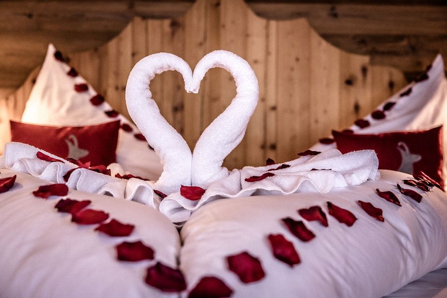 Ein romantisch dekoriertes Bett mit roten Rosenblättern und zwei weißen Handtüchern, die zu einem Herz geformt sind. Der Hintergrund ist aus Holz.
