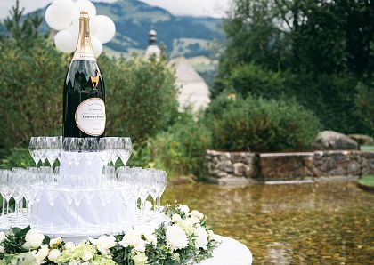 Ein großes Champagnerglas steht auf einer Pyramide aus mehreren Gläsern in einem Garten. Im Hintergrund sind Bäume, Sträucher und eine malerische Landschaft.