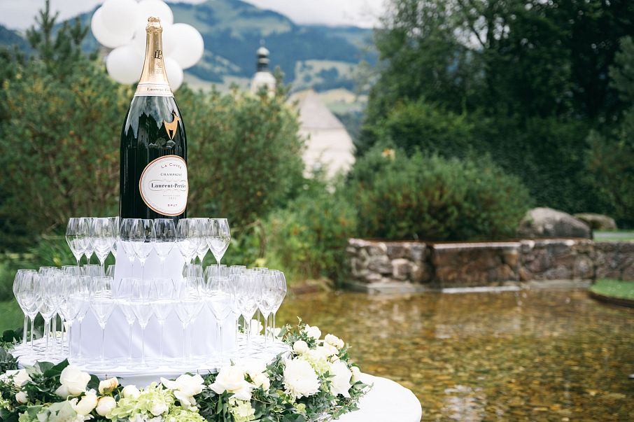 Ein großes Champagnerglas steht auf einer Pyramide aus mehreren Gläsern in einem Garten. Im Hintergrund sind Bäume, Sträucher und eine malerische Landschaft.