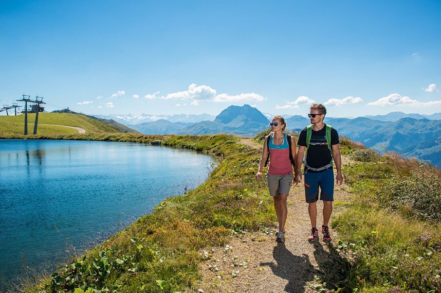 Ein Paar wandert auf einem schmalen Pfad entlang eines Bergsees. Der Himmel ist klar und blau, Berge sind im Hintergrund sichtbar. Beide tragen Rucksäcke.