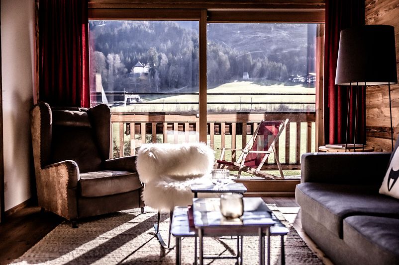 Gemütliches Wohnzimmer mit großen Fenstern, die einen Blick auf die Berge bieten. Ein Sessel, Sofa und Holzmöbel schaffen eine einladende Atmosphäre.