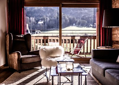 Gemütliches Wohnzimmer mit großen Fenstern, die einen Blick auf die Berge bieten. Ein Sessel, Sofa und Holzmöbel schaffen eine einladende Atmosphäre.